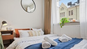 Hotellihuone, jossa on sänky, yöpöytä lampun kanssa, peili, ikkuna verhoilla ja näkymä ulkona oleviin rakennuksiin.