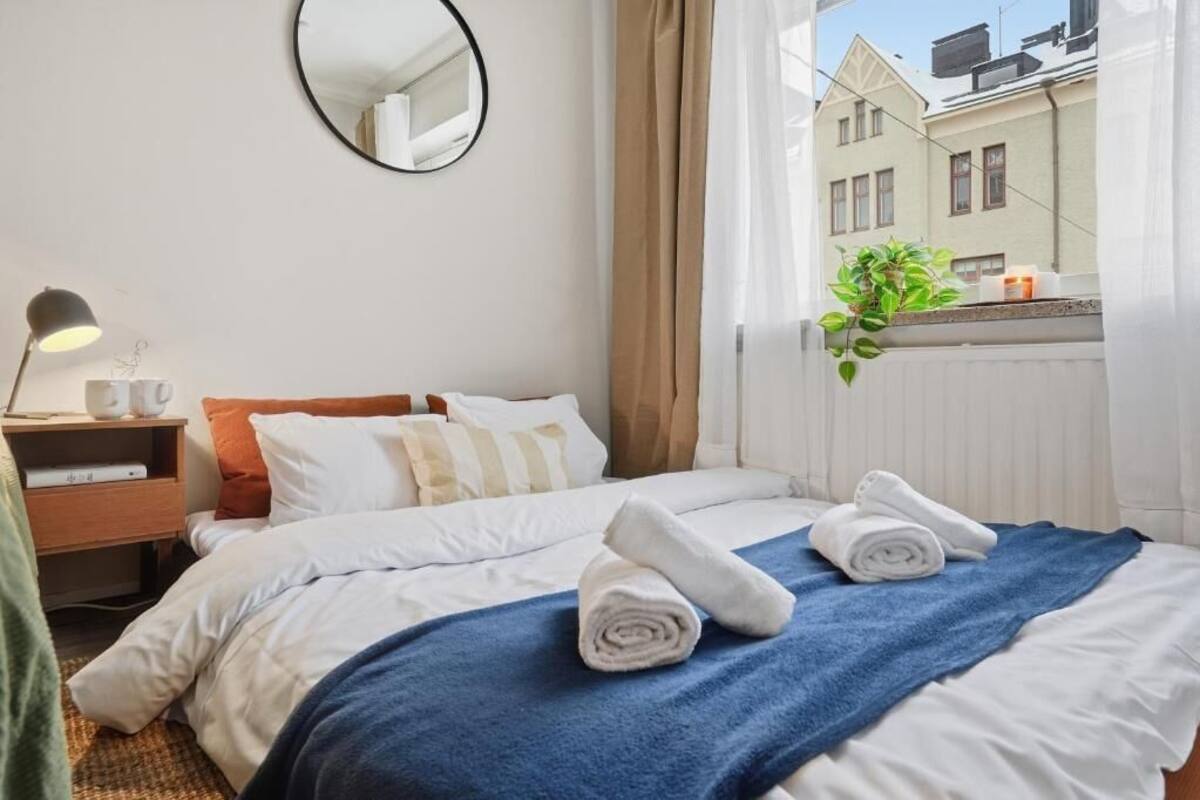 A hotel room with a bed, a bedside table with a lamp, a mirror, a window with curtains, and a view of buildings outside.