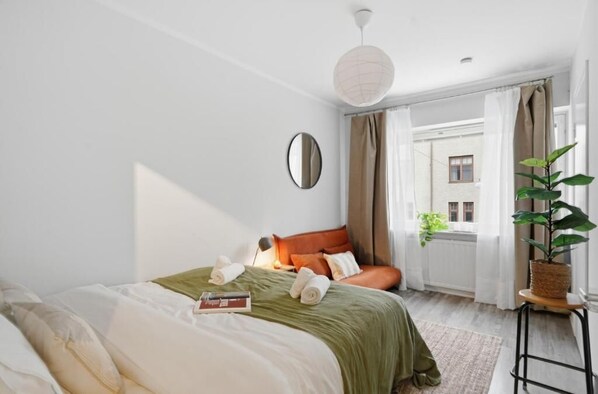 A hotel room with a bed, a bedside table, a mirror, a hanging light fixture, a window with curtains, and a potted plant.
