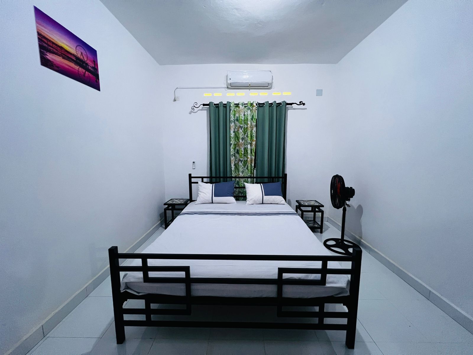 A single bed with a headboard, a bedside table, a wall-mounted air conditioner, a window with curtains, and a wall-mounted fan.
