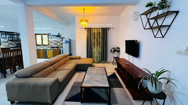 Ein modernes Wohnzimmer mit einem Ecksofa, einem Couchtisch, einem Fernseher und einer im Hintergrund sichtbaren Küche.