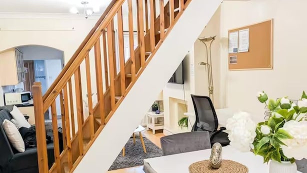 Ein modernes Wohnzimmer mit einer Holztreppe, einem Esstisch mit Stühlen, einer Küchenzeile und einer Pinnwand.