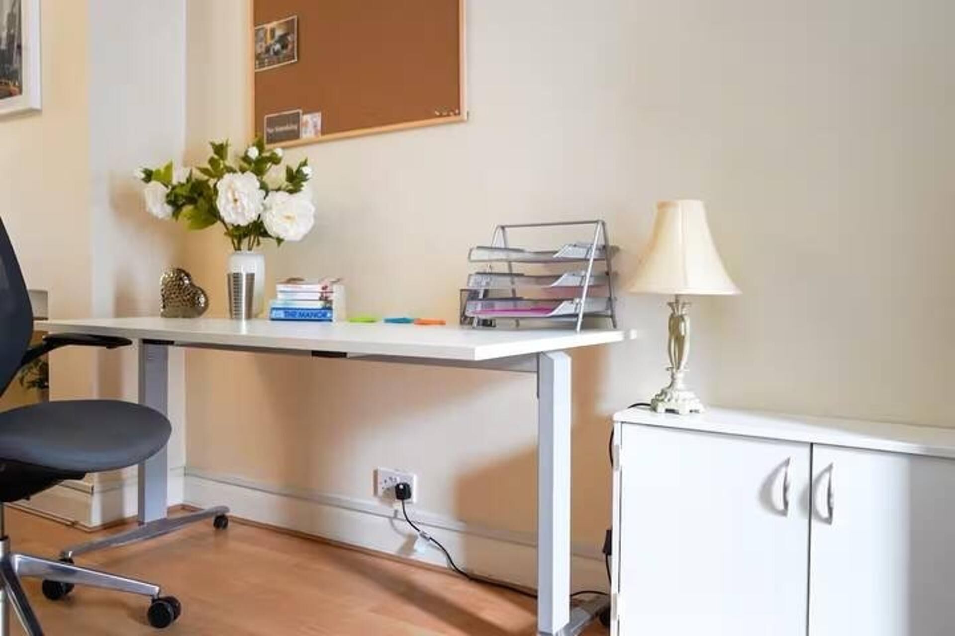 A desk with a lamp, a chair, a cabinet, and a bulletin board with photos.