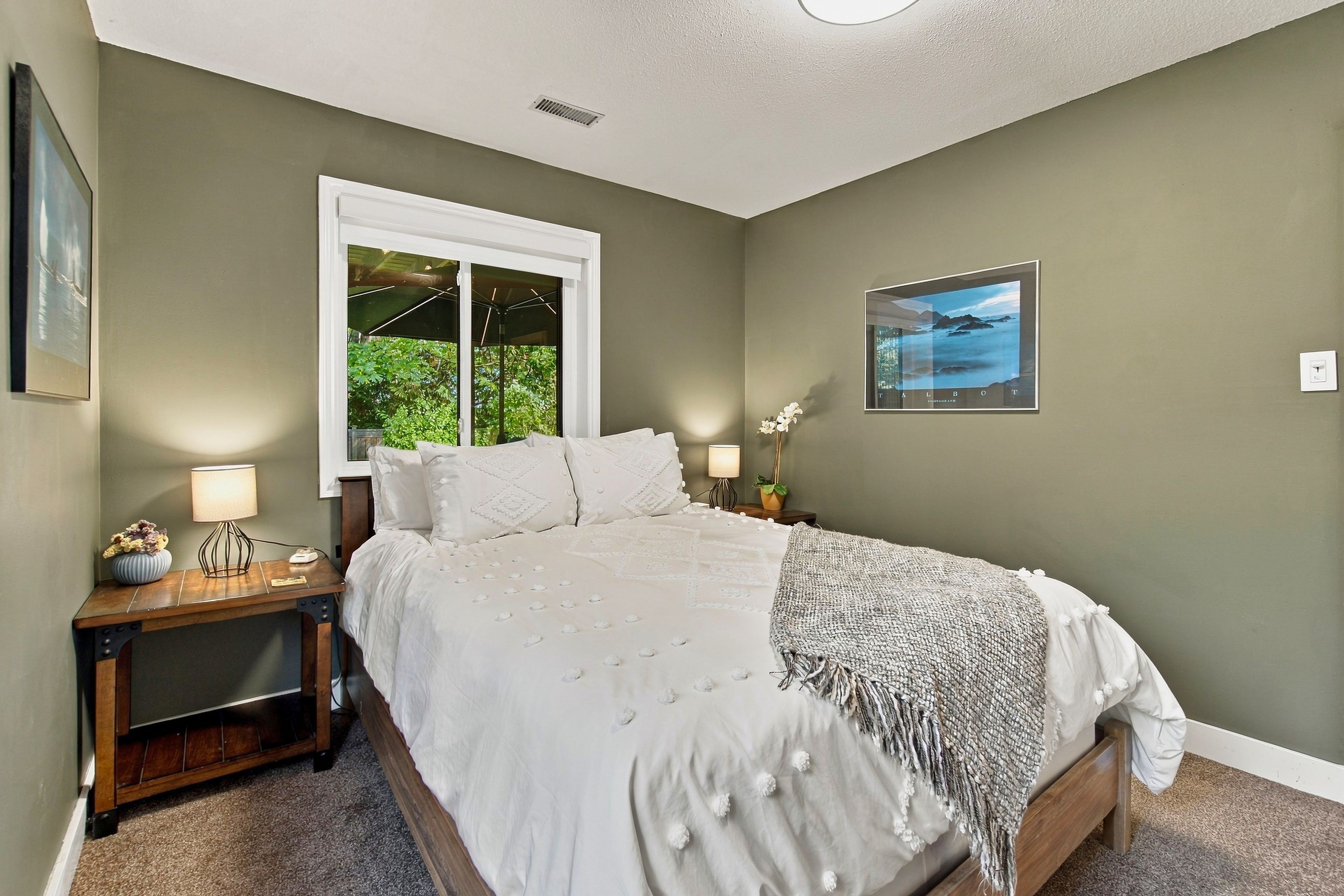 A bedroom with a bed, bedside tables, a window, a wall-mounted picture, and a carpeted floor.