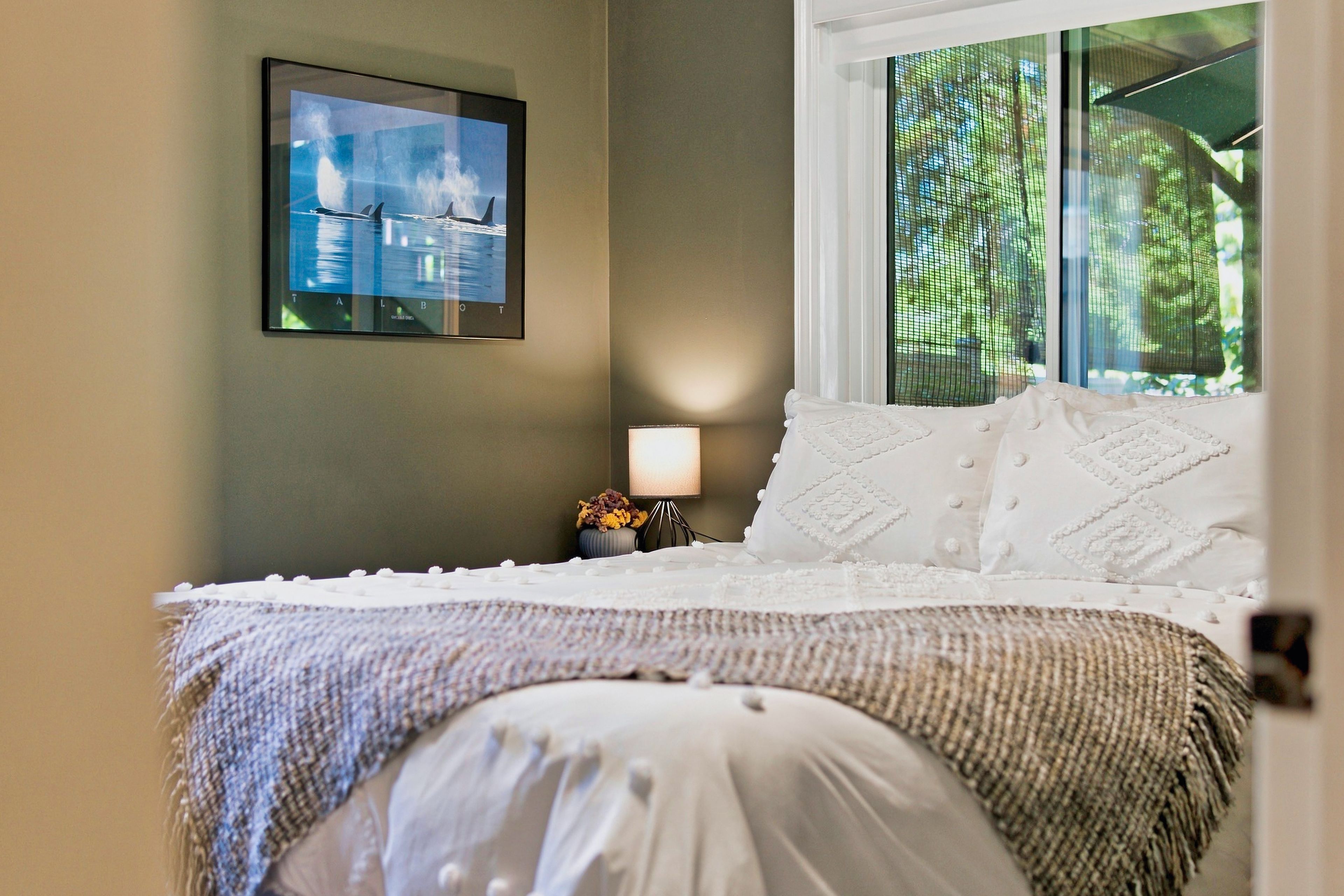 A bedroom with a bed, a nightstand with a lamp, a framed picture on the wall, and a window with a view of trees.
