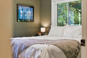 Una camera da letto con un letto, un comodino con una lampada, un quadro appeso al muro e una finestra vista alberi.