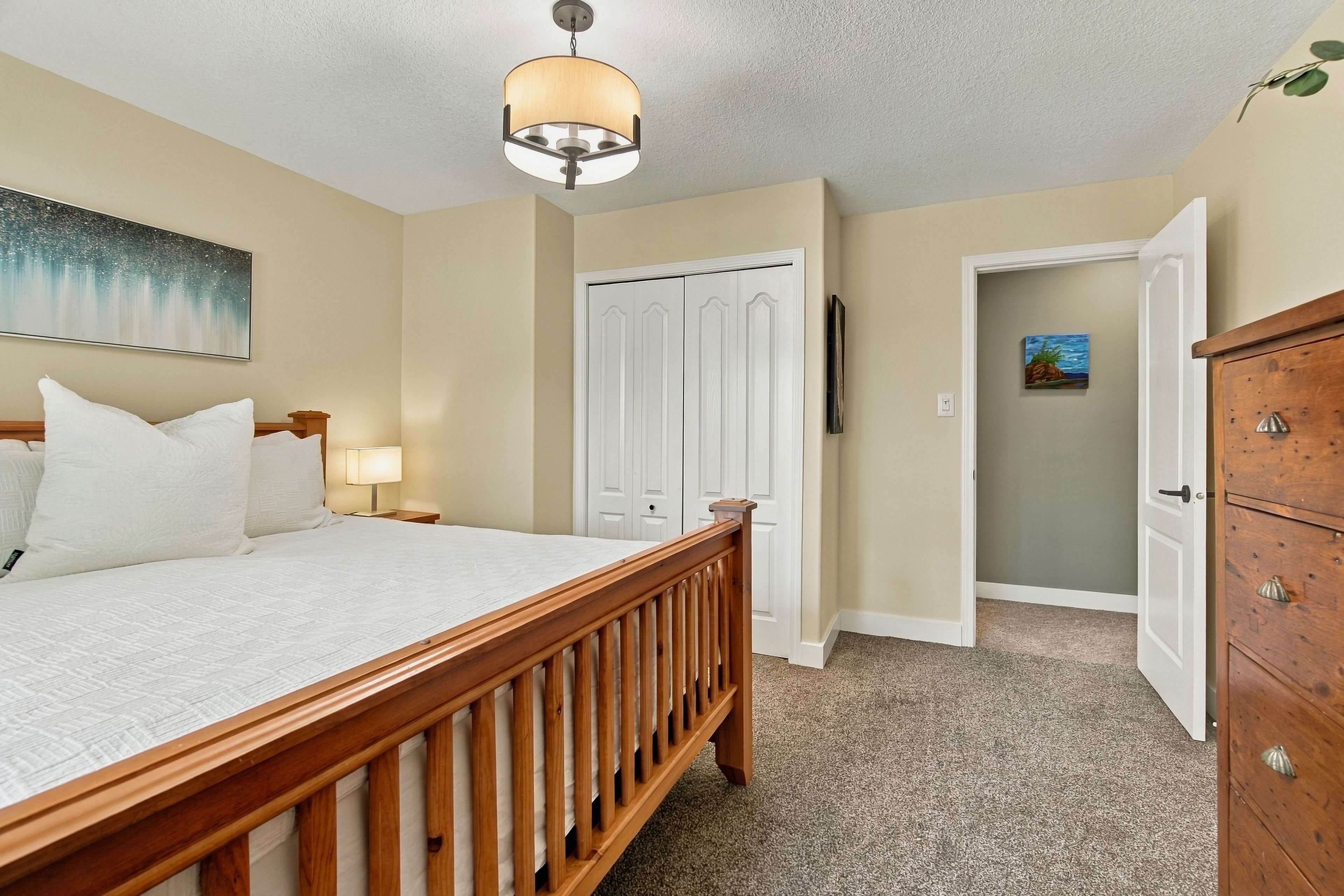 A bedroom with a wooden bed, a nightstand, a lamp, a painting on the wall, and a door leading to another room.