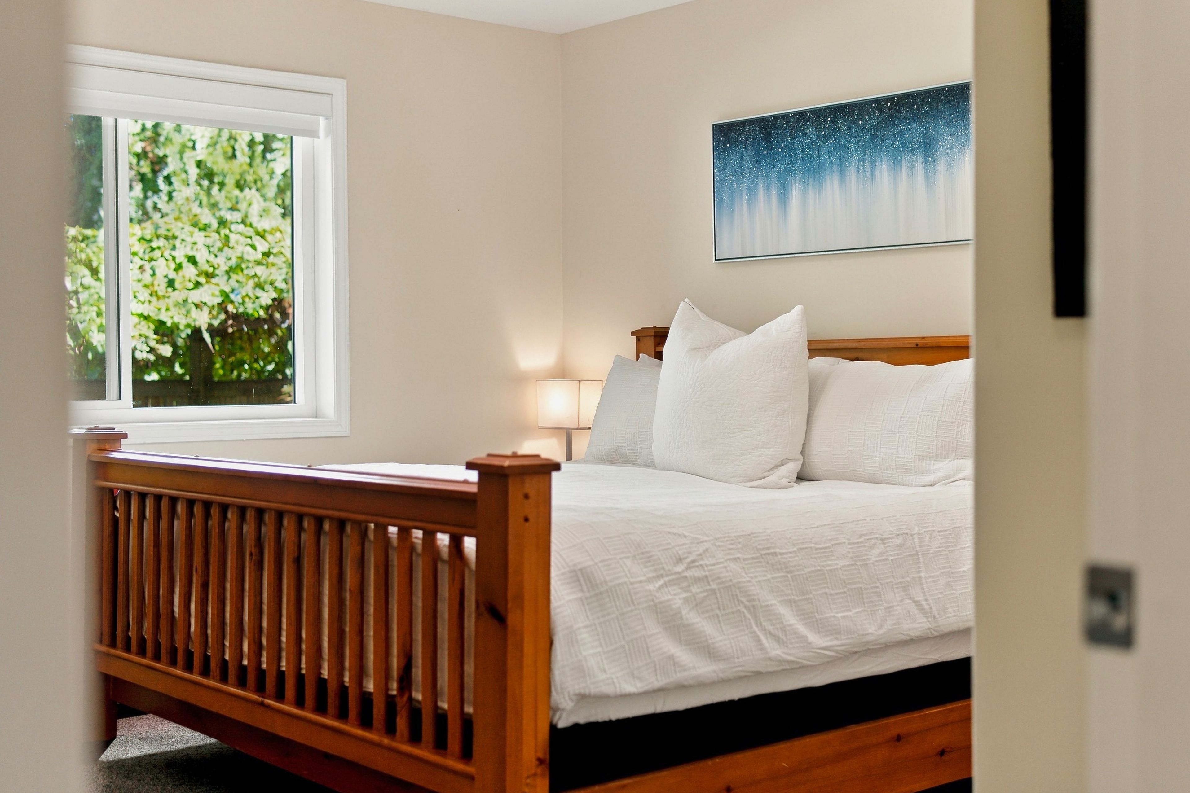 A bedroom with a wooden bed, a window with a view of greenery, and a painting on the wall.