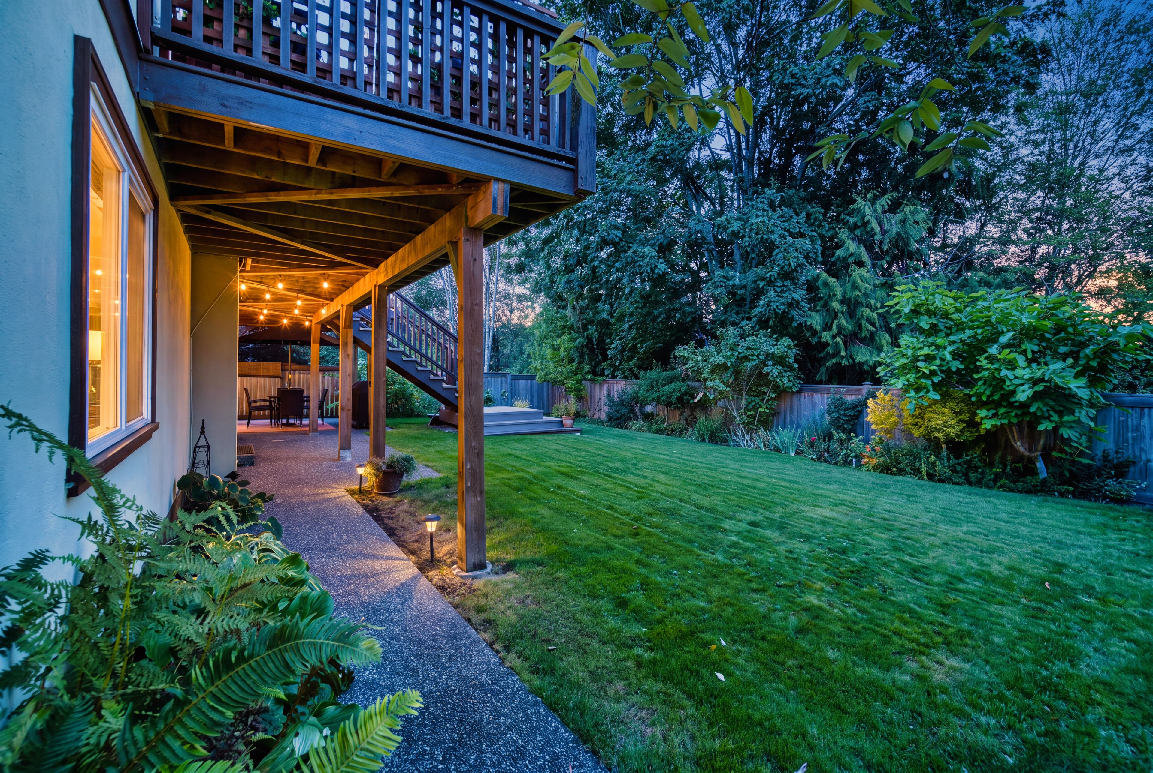 A covered outdoor area with a wooden deck, a staircase leading to a second level, and a well-maintained lawn.