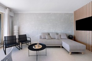 Ein modernes Wohnzimmer mit grauem Sofa, schwarzen Sesseln, einem runden Couchtisch und einem Flachbildfernseher, der an einer Wand aus Holzpaneelen befestigt ist.