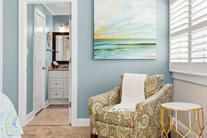 A room with a patterned armchair, a small round table, a painting of a landscape, and a door leading to a bathroom. - Vagabond Penthouse | Ocean Views + Huge Deck (Ocean City)