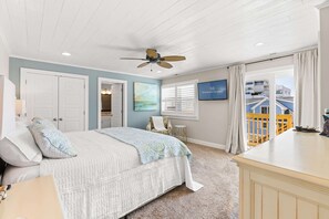 A bedroom with a bed, a ceiling fan, a TV, a window with curtains, and a view of buildings outside. - Vagabond Penthouse | Ocean Views + Huge Deck (Ocean City)