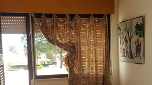 Ein Schlafzimmer mit einem Bett, einem Fenster mit gemusterten Vorhängen, einem an der Wand hängenden Gemälde und einem Korbstuhl.