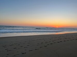 Ein Strand mit Fußspuren, die zum Wasser führen, Wellen und einem Sonnenuntergang.