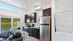 Cocina moderna con refrigerador, microondas y estufa de acero inoxidable, comedor con mesa y sillas, y un amplio ventanal con vista al agua.