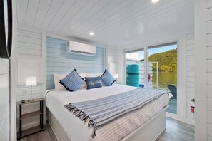 Ein Schlafzimmer mit Bett, Nachttisch, Lampe und Blick auf ein Gewässer durch eine Schiebetür.
