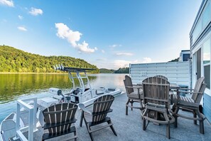 Ein Boot liegt an einem Steg, mit Adirondack-Stühlen und einem Tisch, mit Blick auf einen See vor einer bewaldeten Hügelkulisse.