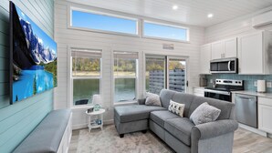 Ein modernes Interieur mit einem grauen Ecksofa, einem Flachbildfernseher, einer Küche mit Edelstahlgeräten und Seeblick durch große Fenster.