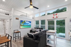 Una habitación amplia con un gran ventanal con vistas a un cuerpo de agua, un ventilador de techo, un cuadro de una escena tropical y un cartel que dice 'Welcome to the Lake'.