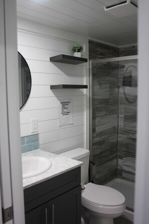 A bathroom with a white sink, dark cabinets, a toilet, a glass shower enclosure, and a potted plant on a shelf. - Driftwood Cove - Pet Friendly Floating House on Lake Cumberland (Russell Springs)