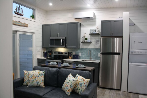 A compact kitchen with grey cabinets, a stainless steel refrigerator, a microwave, and a dining area with a dark sofa and patterned cushions.