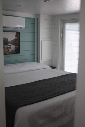 A bedroom with a bed, a window with blinds, and a wall-mounted air conditioner. - Driftwood Cove - Pet Friendly Floating House on Lake Cumberland (Russell Springs)