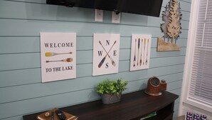 En væg med tre velkomstskilte, en træplante, en vintage radio og et lille bord med en fjernbetjening og en notesbog.