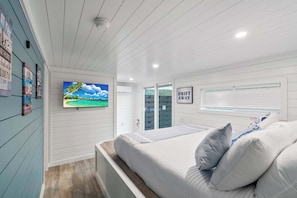 A bedroom with a bed, a television, a window with blinds, and a wall with framed pictures.