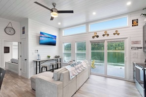 Un intérieur moderne avec un canapé, un ventilateur de plafond, un téléviseur et une vue sur un lac, visible à travers de grandes fenêtres.
