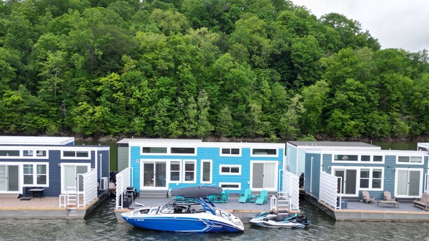 A waterfront property with two blue buildings, a dock with boats, and a forested hill in the background. - Four Suns - Lake Front Floating House Retreat (Russell Springs)