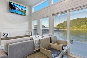 A room with a large window overlooking a lake, a television mounted on the wall, and a sofa with cushions. - On-Water 20 - Pet Friendly Floating House on Lake Cumberland (Russell Springs)
