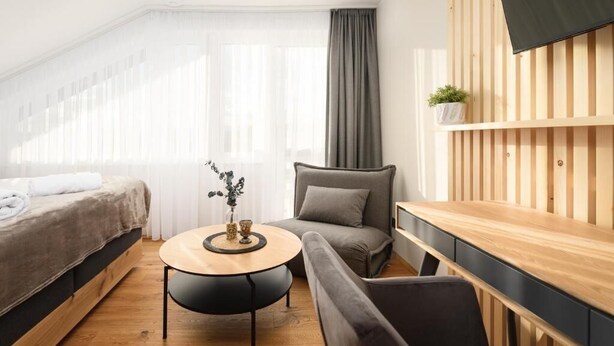 A modern hotel room with a bed, a round table with a vase, a grey sofa, and a wooden TV stand. - Das Weinkitz  (Kitzeck im Sausal)