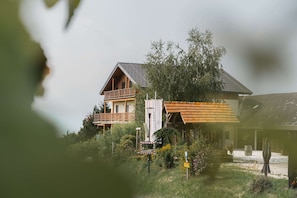 A two-story wooden house with a balcony, surrounded by greenery and a gravel path.