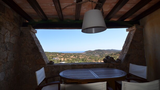 A stone-walled room with a round wooden table and two chairs, a hanging light fixture, and a view of a landscape with hills and a body of water. - Ftam-corn01 in Tamarispa (Budoni)