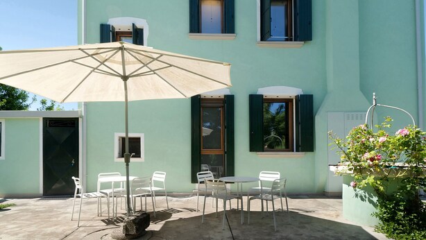 Een gebouw met een lichtgroene gevel, een deur, ramen met luiken, een tafel met stoelen en een parasol.