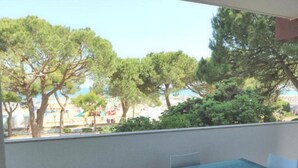 Un balcón con una mesa y sillas de vidrio, con vista a una playa y árboles.