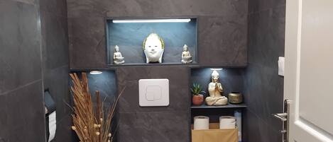 A modern bathroom with dark tiles, a white toilet, a shelf with decorative items, and a small Buddha statue.