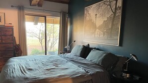 Une chambre à coucher avec un lit, une table de chevet, une lampe, une fenêtre avec des rideaux et un tableau encadré au mur.