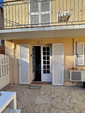 Ein zweigeschossiges Gebäude mit einer Steinterrasse, einer weißen Tür mit Fensterläden und einem Balkon mit Metallgitter.