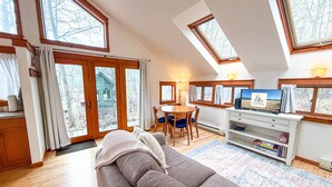 Een ruime woonkamer met een grote bank, een eethoek met een tafel en stoelen, een televisie en een boekenkast. Grote ramen zorgen voor natuurlijk licht en uitzicht op de omgeving.