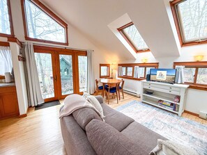 Ein geräumiges Wohnzimmer mit einem großen Sofa, einem Essbereich mit Tisch und Stühlen, einem Fernseher und einem Bücherregal. Große Fenster sorgen für natürliches Licht und Blick ins Freie.