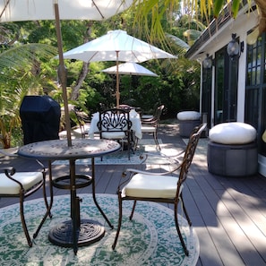 A patio with outdoor furniture, including a table and chairs, under umbrellas, surrounded by lush greenery.