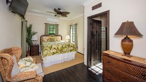 A bedroom with a bed, a television mounted on the wall, a ceiling fan, a door, and a lamp on a side table.
