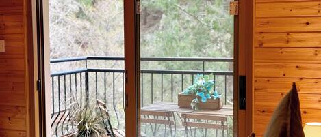 A wooden room with a sliding glass door leading to an outdoor balcony with a table, chairs, and a potted plant.