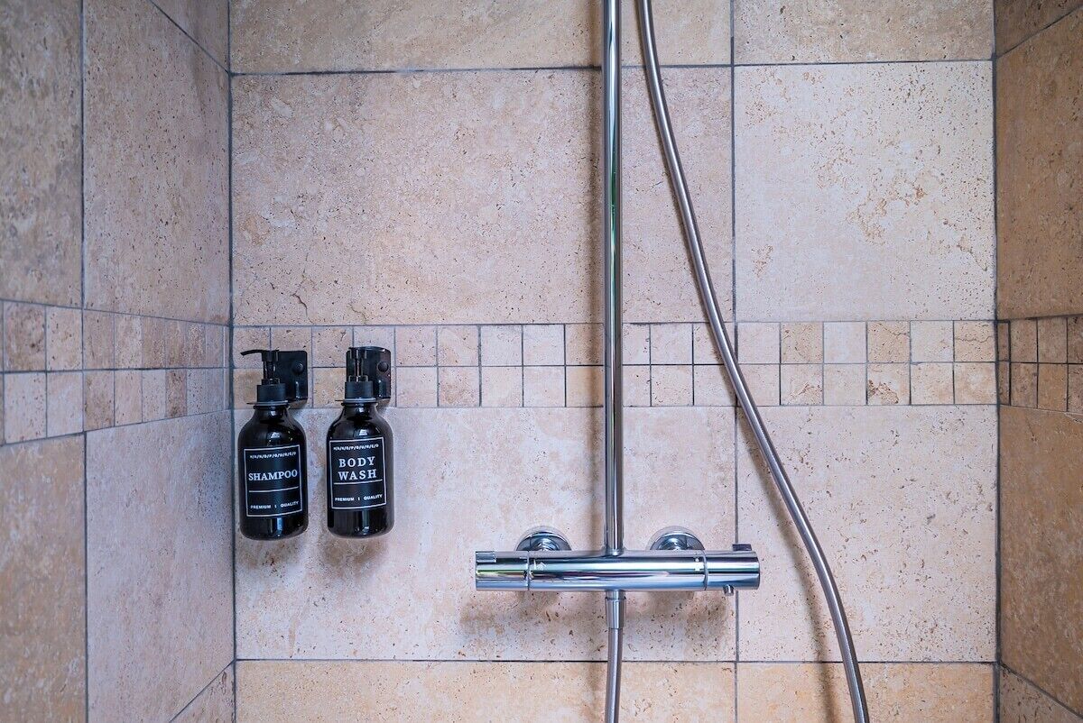 Ein Badezimmer mit beigen Fliesen, einem chromfarbenen Duschkopf und zwei schwarzen Flaschen, beschriftet mit 'shampoo' und 'body wash', an der Wand befestigt.