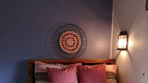 Una cama con una colcha estampada, dos almohadas, un adorno de pared y una lámpara fijada a la pared.