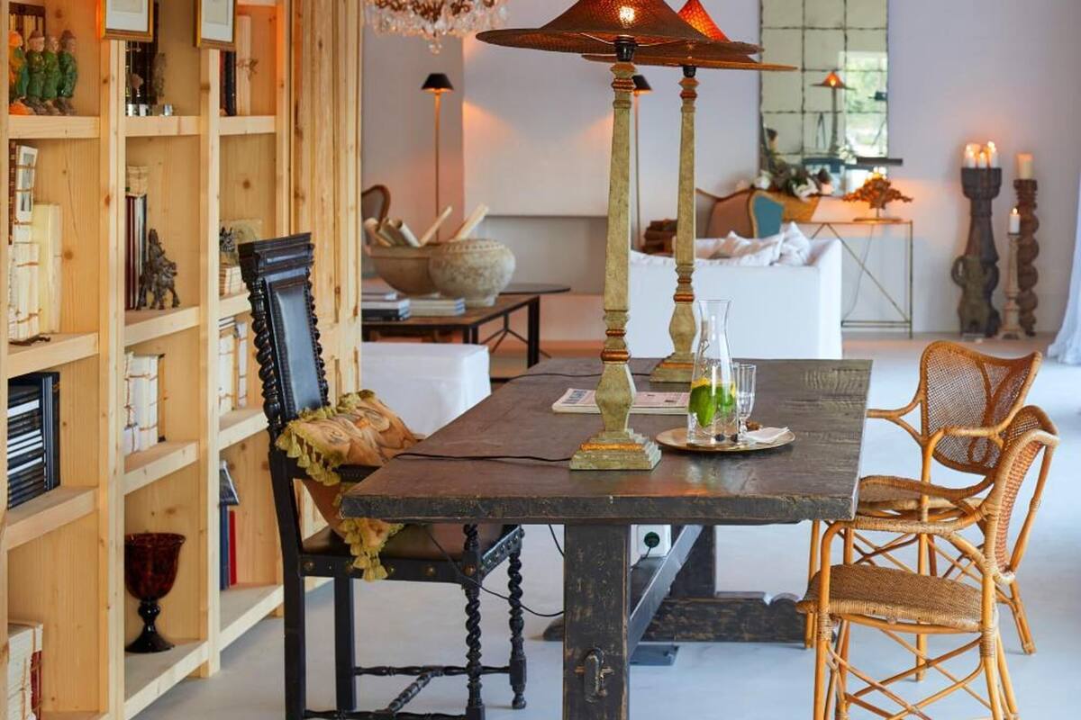 A dining area with a wooden table, chairs, and a chandelier. There is a bookshelf filled with various items and a mirror on the wall.