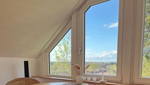 Een kamer met een houten tafel, rieten stoelen, grote ramen met uitzicht op bomen en gebouwen, en een witte muur.