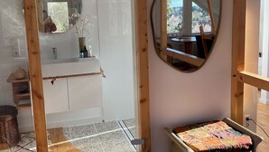 Een kamer met een houten balkenplafond, een ronde spiegel, een klein tafeltje met een kleurrijke quilt, en uitzicht op een tuin door een raam.