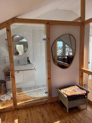 A room with a wooden ceiling structure, a round mirror, a small table with a colorful quilt, and a view of a garden through a window. - Cozy attic studio on the Lech. Nature and old town flair in Landsberg (Landsberg am Lech)